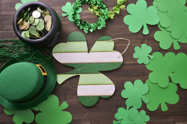 Overhead View of an Assortment of St. Patrick’s Day Decorations – Painted Wood Shamrock, Leprechaun Hat, Green Clover Clings and a Pot of Gold on a Dark Wood Background