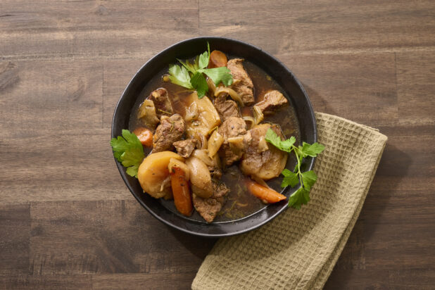 Close Up Overhead View of a Large Black Bowl of Irish Stew With Carrots, Potatoes and Beef on a Dark Wooden Table with a Beige Napkin