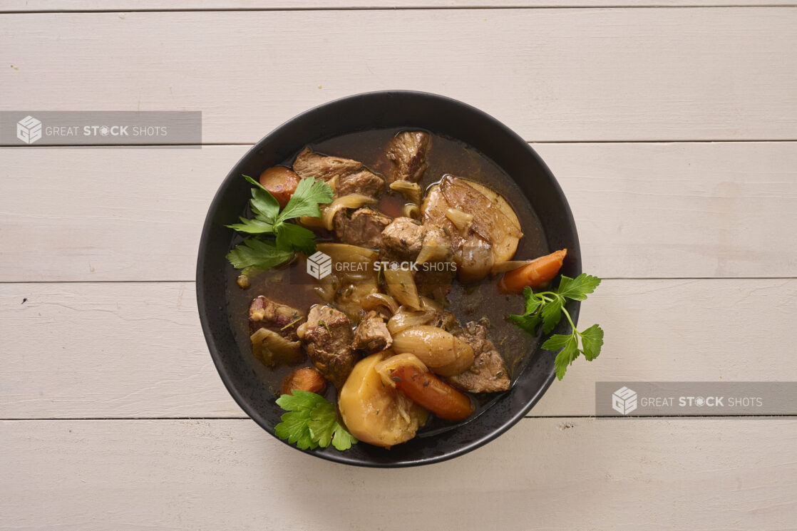 Close Up Overhead View of a Large Black Bowl of Irish Stew With Carrots, Potatoes and Beef on a White Painted Wood Table