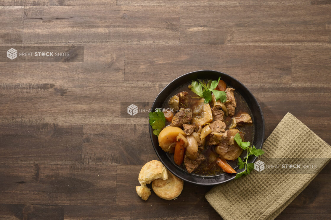 Overhead View of a Large Black Bowl of Irish Stew With Carrots, Potatoes and Beef on a Dark Wooden Table with Butter Biscuits and a Beige Napkin