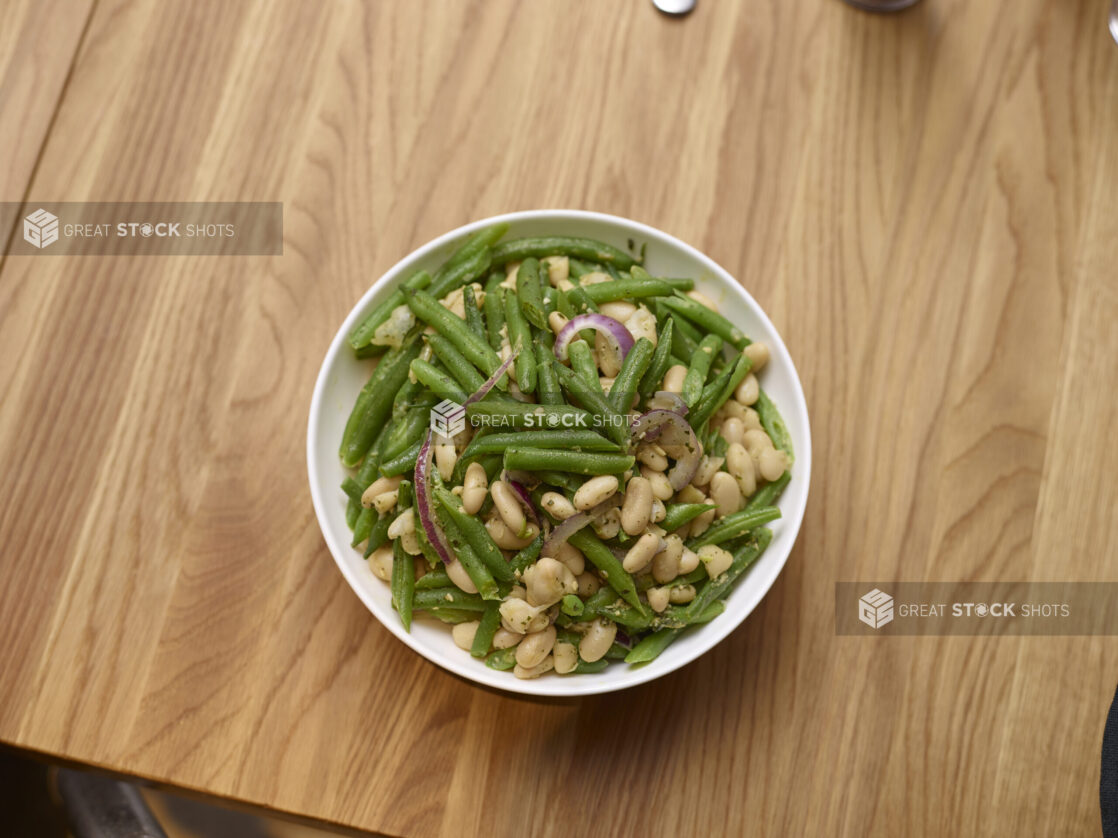 Overhead View of a Cold Italian Green Bean Salad with White Beans, Sliced Red Onions in a White Ceramic Salad Bowl on a Wooden Surface