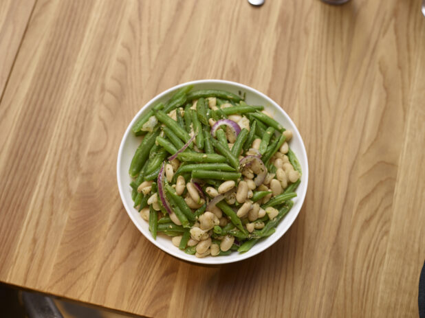 Overhead View of a Cold Italian Green Bean Salad with White Beans, Sliced Red Onions in a White Ceramic Salad Bowl on a Wooden Surface