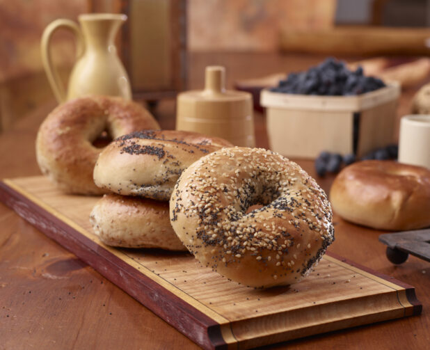 Various whole bagels on a wooden board in the foreground with a basket of blueberries and other accessories in the background