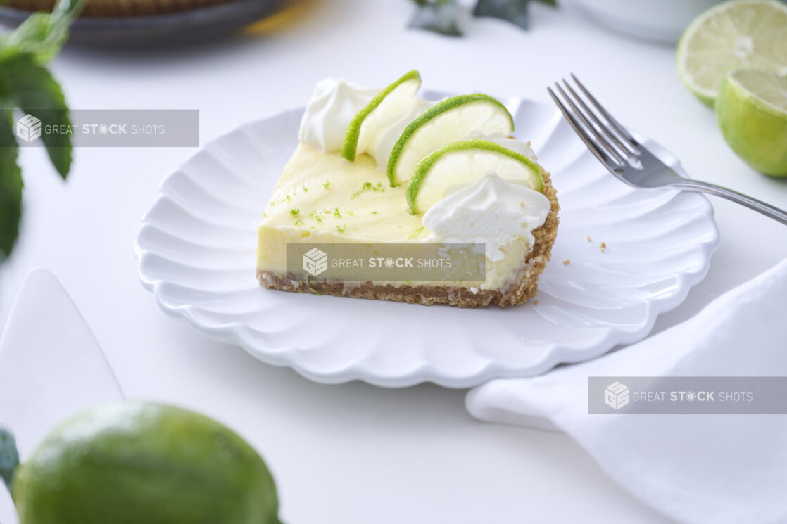 Close Up of a Slice of Key Lime Pie with Whipped Cream and Lime Slice Garnish on a Decorative White Plate on a White Table Surface in an Indoor Setting