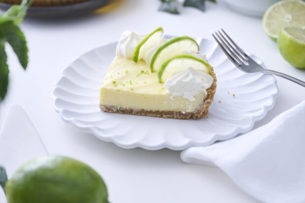 Close Up of a Slice of Key Lime Pie with Whipped Cream and Lime Slice Garnish on a Decorative White Plate on a White Table Surface in an Indoor Setting
