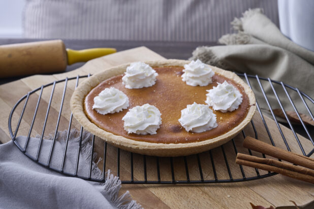 A Whole Pumpkin Pie with Whipped Cream Garnish and Cinnamon Sticks on a Cooling Rack in an Indoor Setting