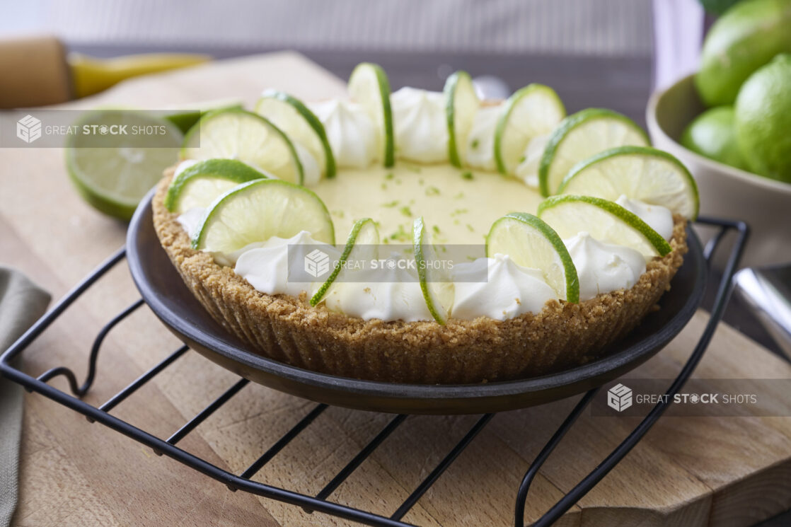 A Whole Key Lime Pie with Sliced Lime and Whipped Cream Garnish on a Cooling Rack in an Indoor Setting