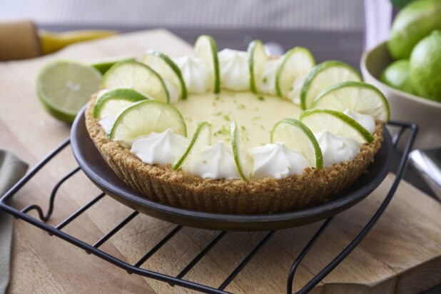 A Whole Key Lime Pie with Sliced Lime and Whipped Cream Garnish on a Cooling Rack in an Indoor Setting