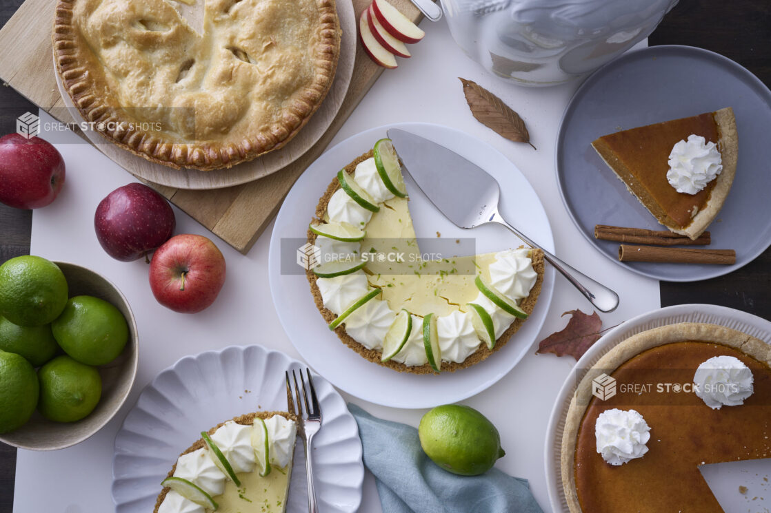 Overhead View of a Group of Fruit Pies (Apple, Key Lime, Pumpkin) with Fresh Apples and Limes on a White Table Surface