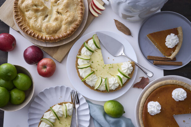 Overhead View of a Group of Fruit Pies (Apple, Key Lime, Pumpkin) with Fresh Apples and Limes on a White Table Surface