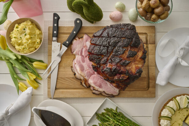 Overhead View of an Easter Ham Roast on a Wooden Cutting Board with Mashed Potatoes and Other Accompaniments, on a White Wooden Table - Variarion