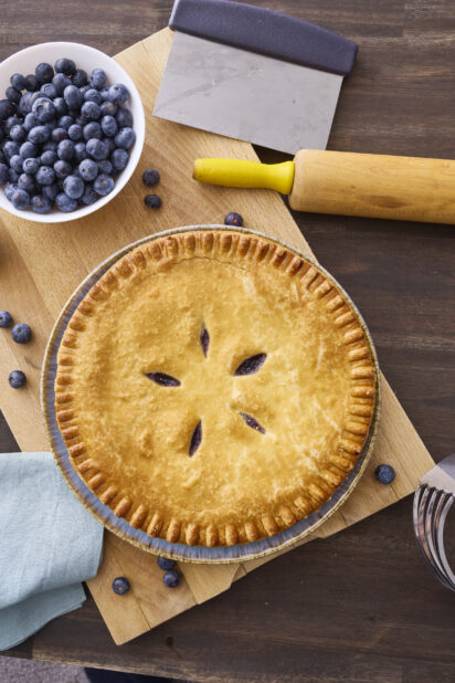 Overhead View of a Whole Blueberry Pie with a Bowl of Fresh Blueberries on a Wooden Cutting Board in an Indoor Setting