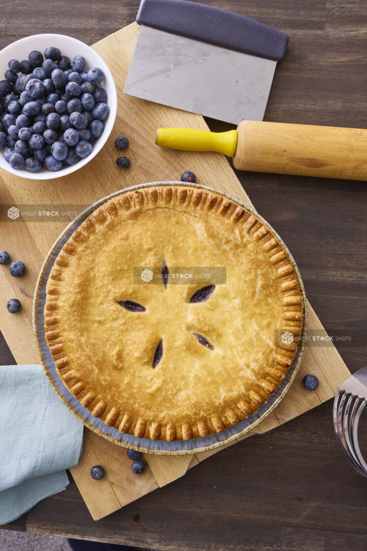 Overhead View of a Whole Blueberry Pie with a Bowl of Fresh Blueberries on a Wooden Cutting Board in an Indoor Setting