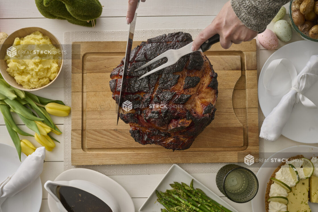 Overhead View of Hands Carving an Easter Ham Roast on a Wooden Cutting Board with Mashed Potatoes and Other Sides, on a White Wooden Table