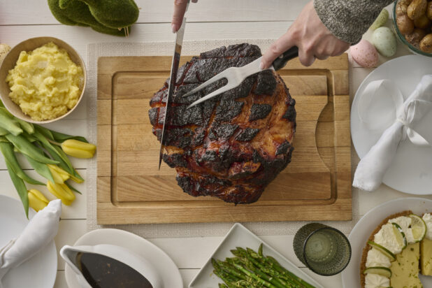 Overhead View of Hands Carving an Easter Ham Roast on a Wooden Cutting Board with Mashed Potatoes and Other Sides, on a White Wooden Table