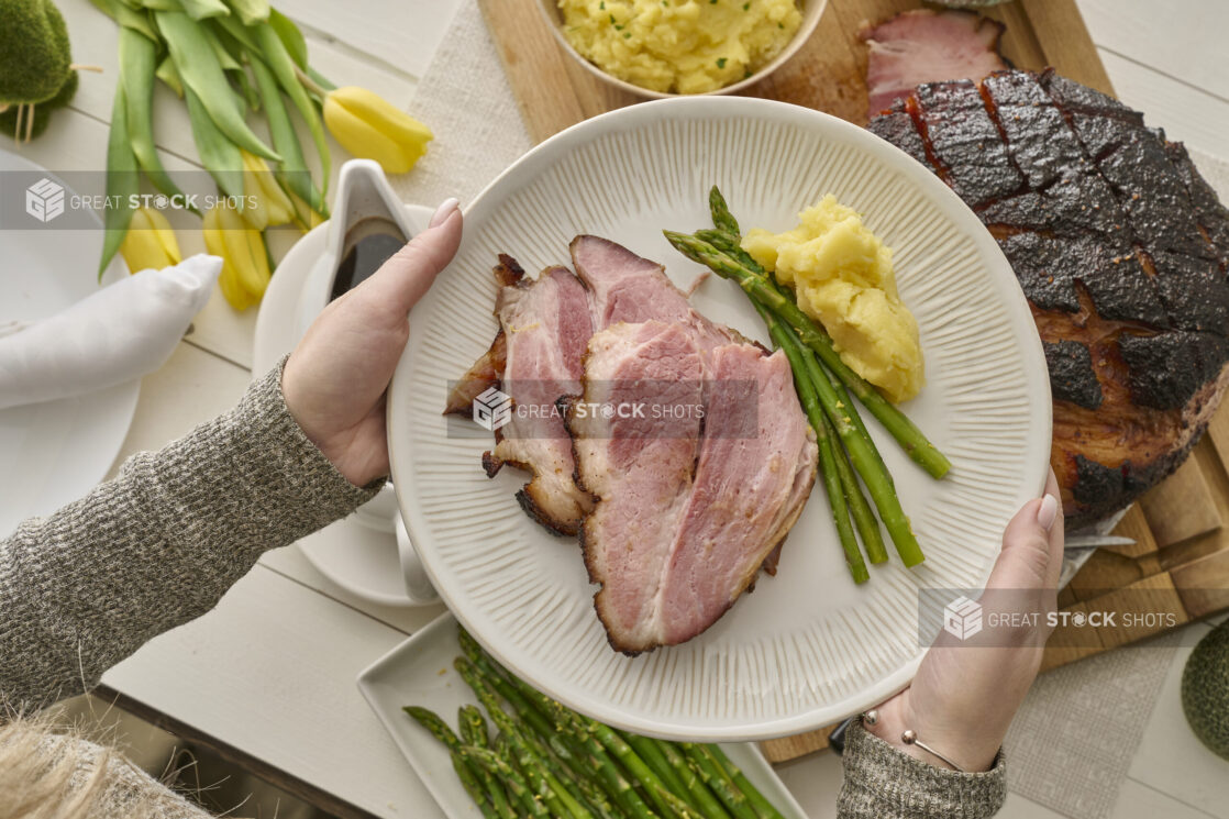 Overhead View of a Person Holding a Plate of Food for Easter Dinner (Easter Ham, Asparagus, Mashed Potatoes) in an Indoor Family Dinner Setting