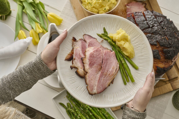 Overhead View of a Person Holding a Plate of Food for Easter Dinner (Easter Ham, Asparagus, Mashed Potatoes) in an Indoor Family Dinner Setting