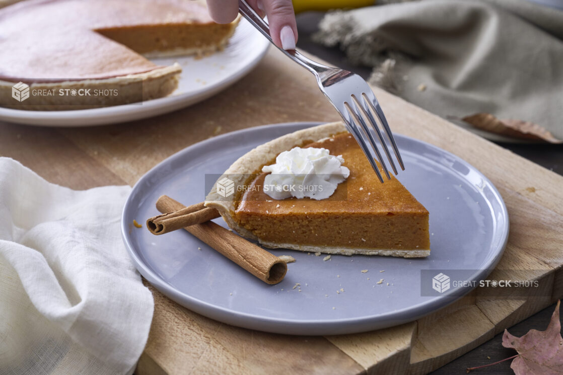 Person with a Fork About to Eat a Slice of Pumpkin Pie with Whipped Cream Garnish and Cinnamon Sticks on a Grey Ceramic Dish