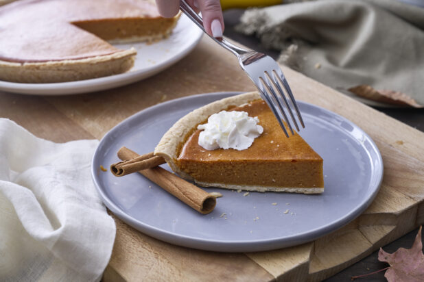 Person with a Fork About to Eat a Slice of Pumpkin Pie with Whipped Cream Garnish and Cinnamon Sticks on a Grey Ceramic Dish