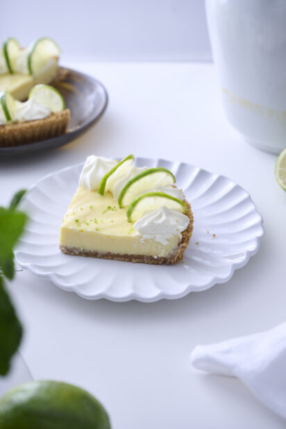 Slice of Key Lime Pie with Whipped Cream and Lime Slice Garnish on a Decorative White Plate on a White Table Surface in an Indoor Setting