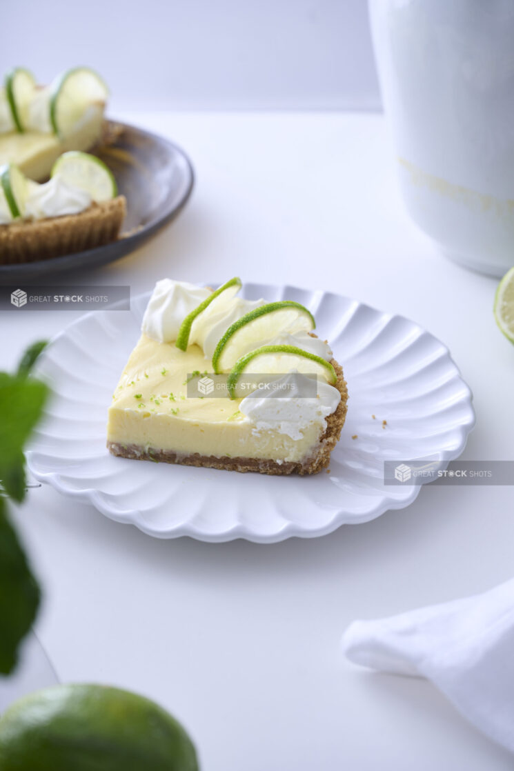 Slice of Key Lime Pie with Whipped Cream and Lime Slice Garnish on a Decorative White Plate on a White Table Surface in an Indoor Setting