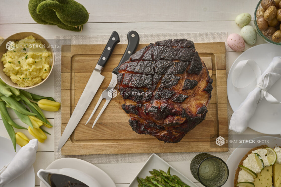 Overhead View of an Easter Ham Roast on a Wooden Cutting Board with Mashed Potatoes and Other Accompaniments, on a White Wooden Table