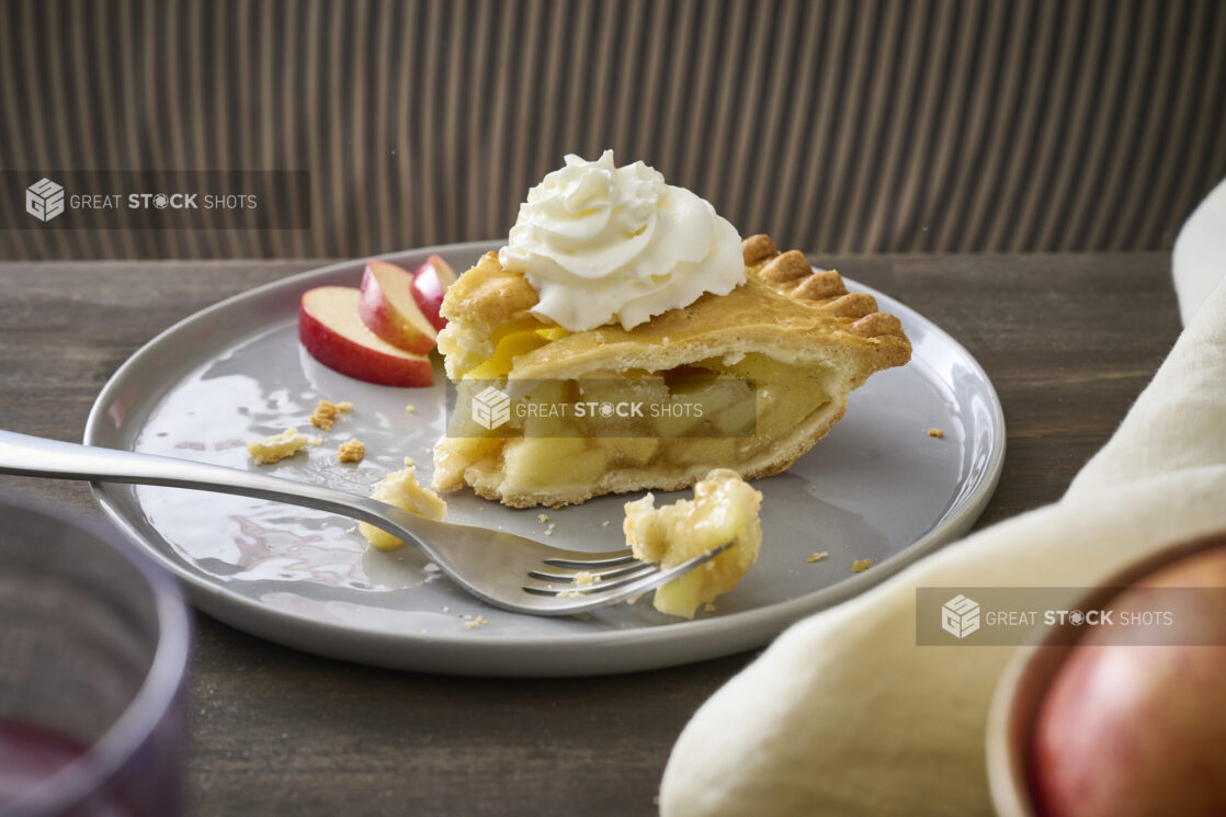 Partially-Eaten Apple Pie Slice with Whipped Cream Garnish and Sliced Apples on a Grey Ceramic Dish