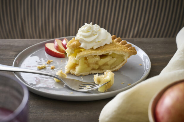 Partially-Eaten Apple Pie Slice with Whipped Cream Garnish and Sliced Apples on a Grey Ceramic Dish