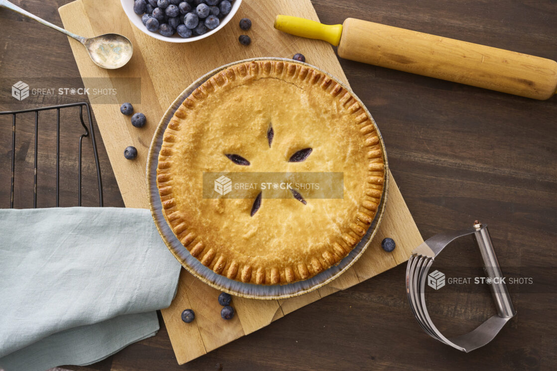 Overhead View of a Whole Blueberry Pie with a Bowl of Fresh Blueberries on a Wooden Cutting Board in an Indoor Setting - Variation