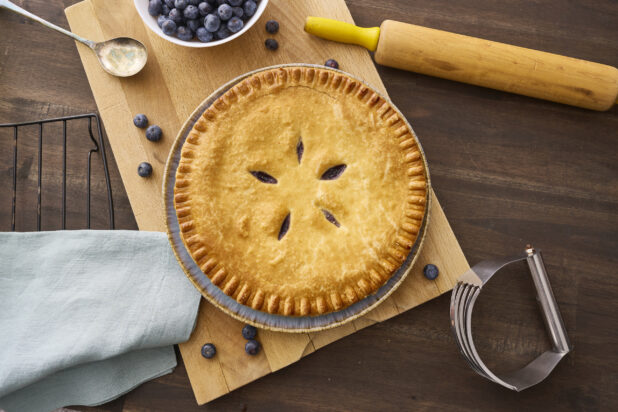 Overhead View of a Whole Blueberry Pie with a Bowl of Fresh Blueberries on a Wooden Cutting Board in an Indoor Setting - Variation