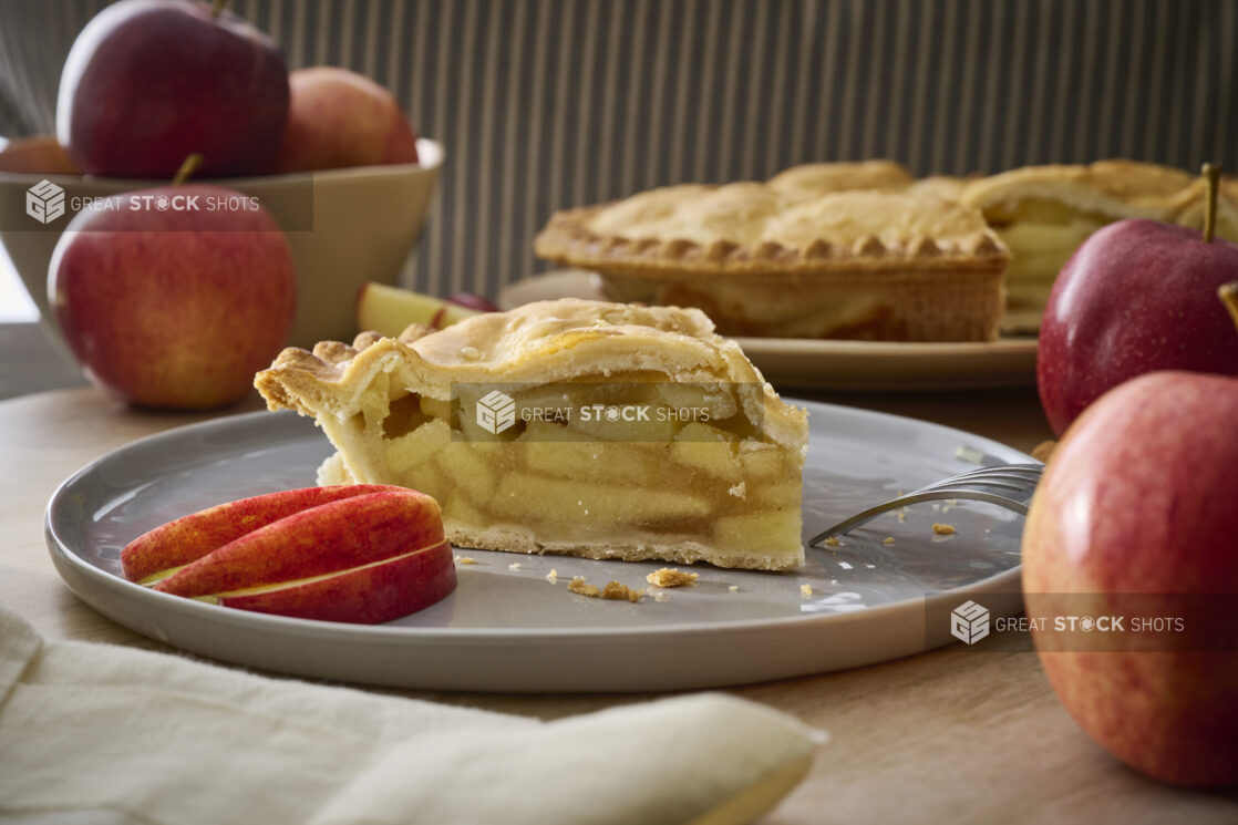 Slice of Apple Pie with Fresh Apple Slices on a Grey Ceramic Dish Surrounded by Fresh Whole Apples in an Indoor Setting