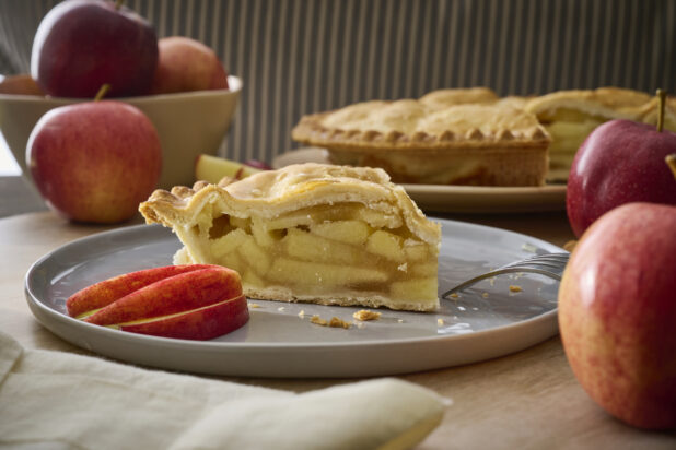 Slice of Apple Pie with Fresh Apple Slices on a Grey Ceramic Dish Surrounded by Fresh Whole Apples in an Indoor Setting
