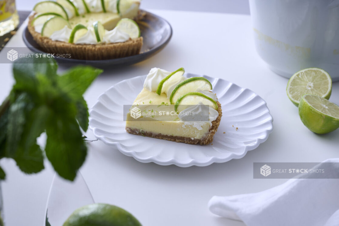 Slice of Key Lime Pie with Whipped Cream and Lime Slice Garnish on a Decorative White Plate on a White Table Surface in an Indoor Setting - Variation