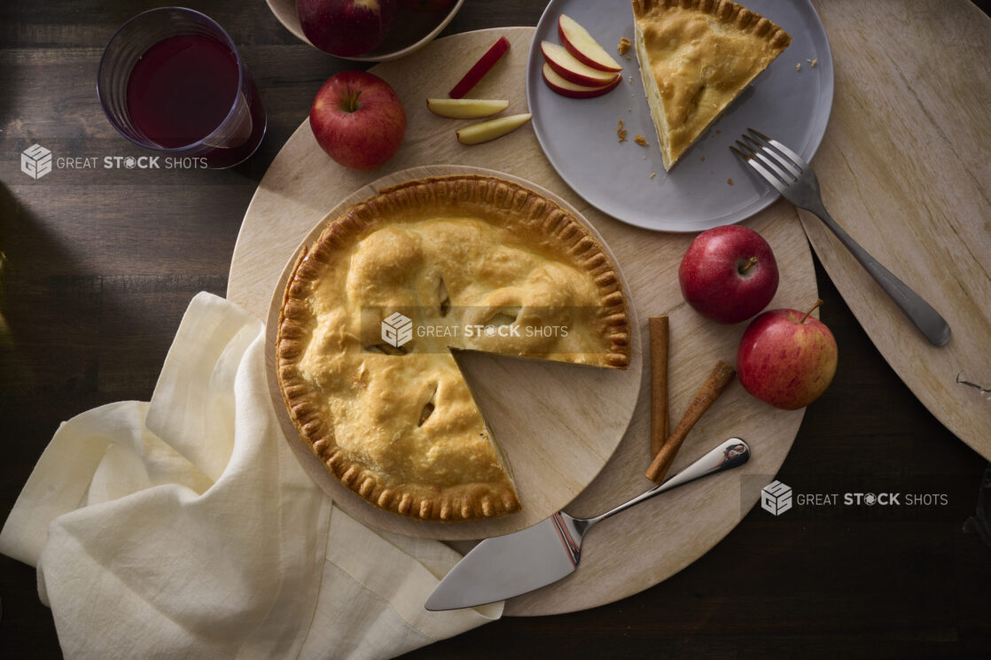 Overhead View of an Apple Pie With a Slice Taken Out, Surrounded by Whole Fresh Apples on a Wooden Table Surface