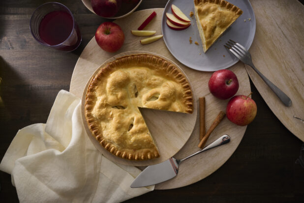 Overhead View of an Apple Pie With a Slice Taken Out, Surrounded by Whole Fresh Apples on a Wooden Table Surface