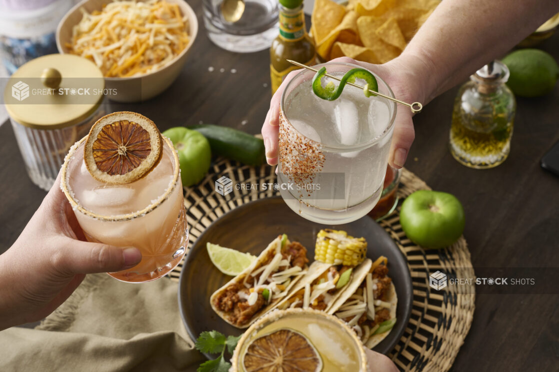 Hands Holding Assorted Citrus Margaritas Doing Cheers Over a Table of Mexican Food and Snacks in an Indoor Setting - Sequence 7