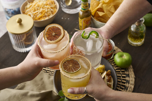 Hands Holding Assorted Citrus Margaritas Doing Cheers Over a Table of Mexican Food and Snacks in an Indoor Setting - Sequence 2