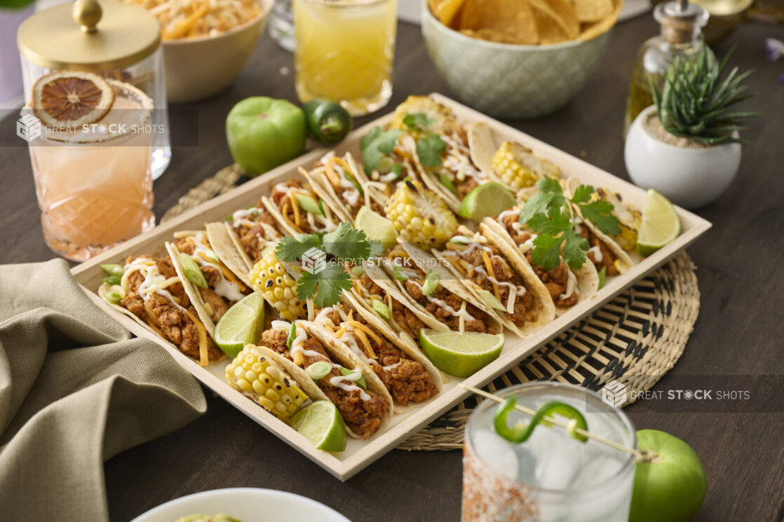 Close-Up of a Wood Catering Platter of Soft Tacos and Roasted Corn on a Woven Placemat, Surrounded by Citrus Margaritas on a Dark Wood Table in an Indoor Setting