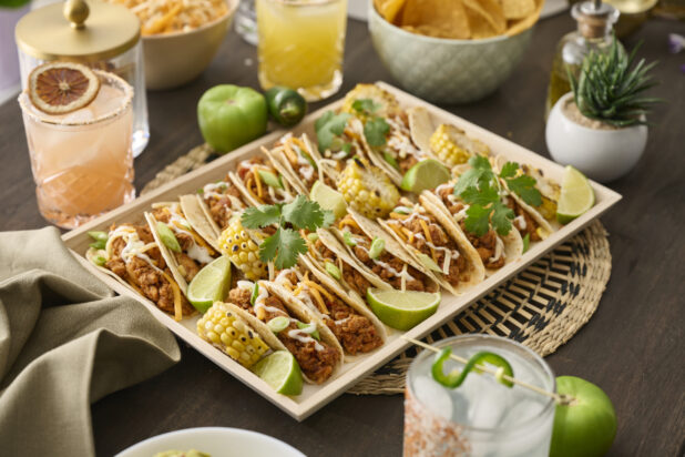 Close-Up of a Wood Catering Platter of Soft Tacos and Roasted Corn on a Woven Placemat, Surrounded by Citrus Margaritas on a Dark Wood Table in an Indoor Setting