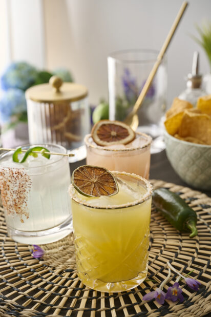 Orange Margarita with a Dried Lime Wheel and Other Assorted Margaritas in Glass Tumblers on a Woven Placemat in an Indoor Setting