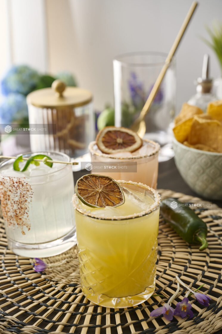 Orange Margarita with a Dried Lime Wheel and Other Assorted Margaritas in Glass Tumblers on a Woven Placemat in an Indoor Setting