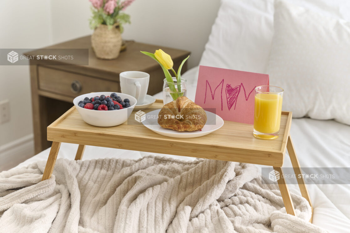 Breakfast in Bed From a Child to Their Mother with Croissant and Berries on a Bamboo Tray Table in a Bedroom Setting