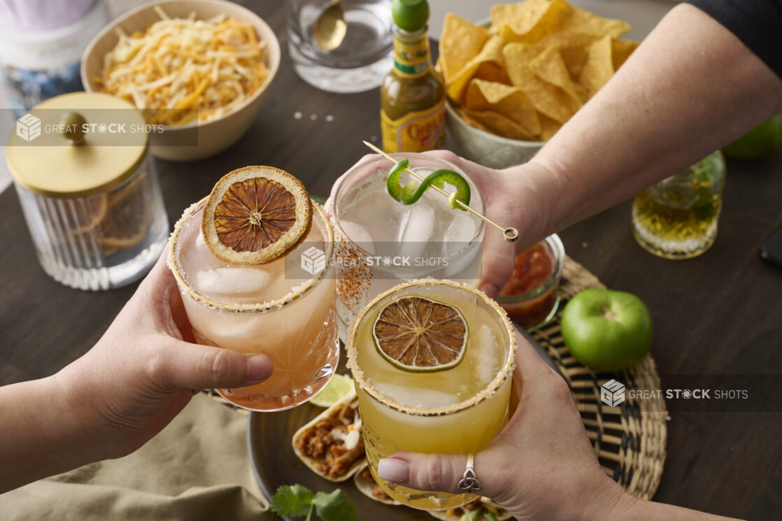 Hands Holding Assorted Citrus Margaritas Doing Cheers Over a Table of Mexican Food and Snacks in an Indoor Setting - Sequence 1