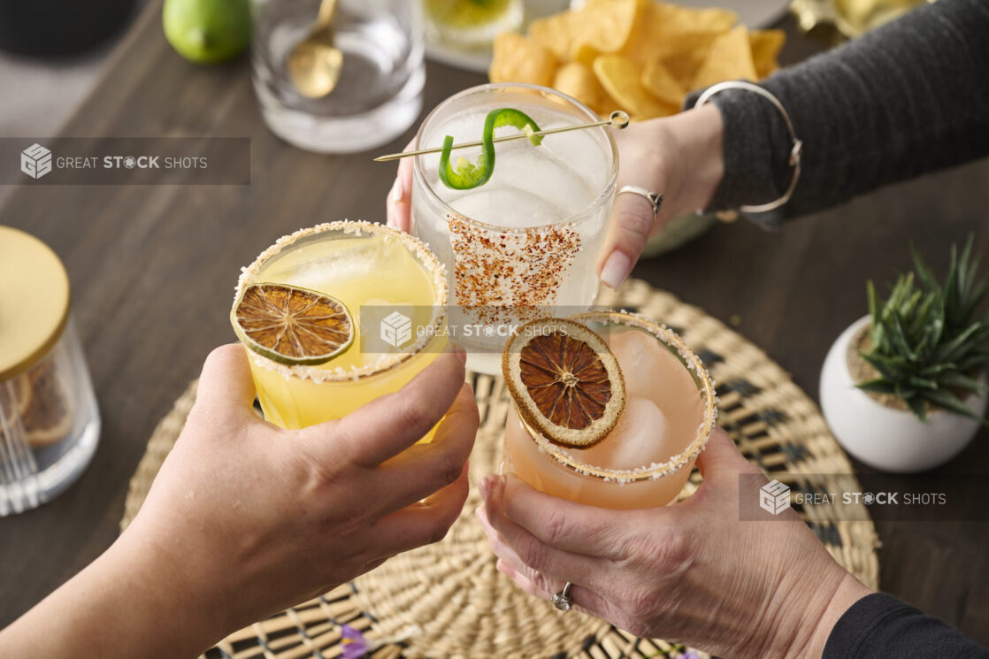 Hands Holding a Trio of Colourful Margarita Cocktails Doing Cheers in an Indoor Setting