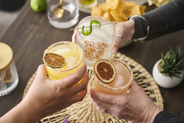 Hands Holding a Trio of Colourful Margarita Cocktails Doing Cheers in an Indoor Setting