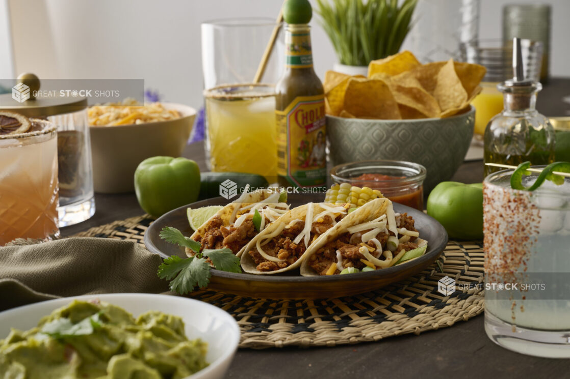 A Trio of Soft Tacos and Roasted Corn on the Cob in a Dark Brown Dish on a Woven Placemat in an Indoor Setting - Variation