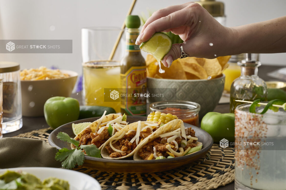 A Woman’s Hand Squeezes a Fresh Lime Wedge Over a Trio of Soft Tacos in a Dark Brown Dish in an Indoor Setting – Sequence 2