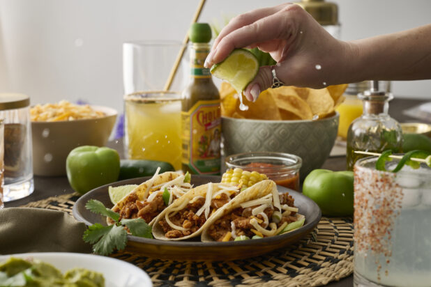 A Woman’s Hand Squeezes a Fresh Lime Wedge Over a Trio of Soft Tacos in a Dark Brown Dish in an Indoor Setting – Sequence 2