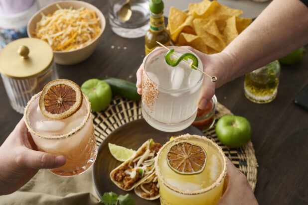 Hands Holding Assorted Citrus Margaritas Doing Cheers Over a Table of Mexican Food and Snacks in an Indoor Setting - Sequence 6
