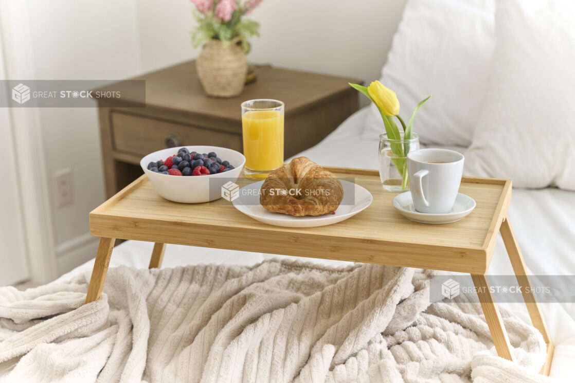 Breakfast in Bed with Croissant, a Bowl of Berries, Coffee and Juice on a Bamboo Tray Table in a Bedroom Setting